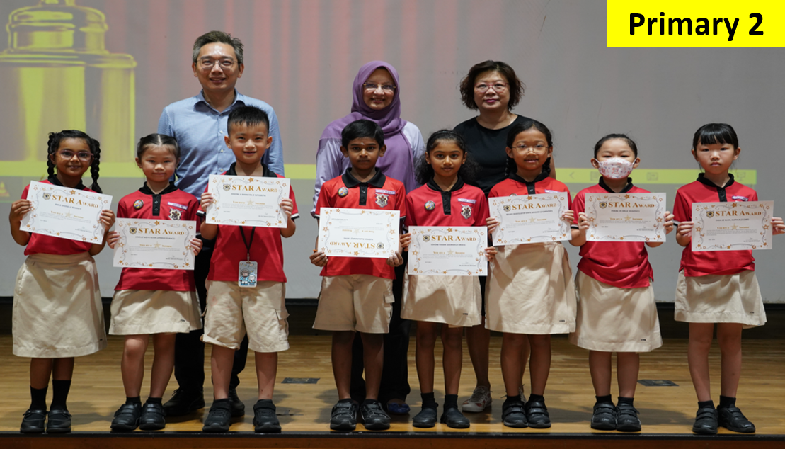 Seven Primary 2 students in red uniforms hold Star Award certificates, posed with three adults on a stage.